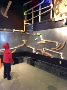 A young boy in a red sweatshirt looking at a wall with tubes and pipes.