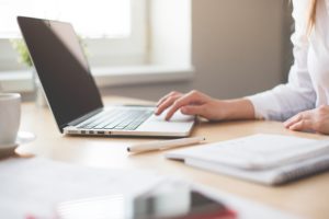 Woman typing on a laptop, a notebook and pen are in the foreground.