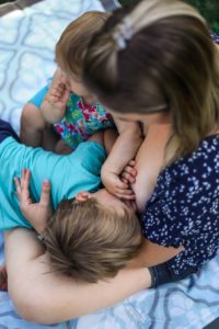 Mother with brown hair and blue flowered shirt nurses older toddler while younger sibling sucks her thumb.