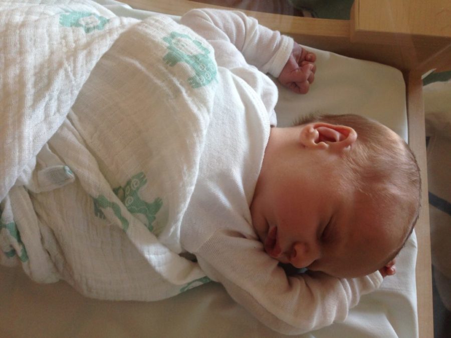 A Pediatric Nurse and Doula’s Initial Breastfeeding Journey Baby wearing white long sleeve shirt sleeping on their back, arms above their head, wrapped in a white swaddle blanket with teal lions on it.