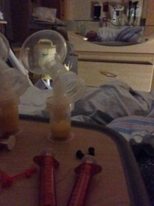 Image of hospital room table with pumped breastmilk and feeding syringes. Infant swaddled in bassinet in background.