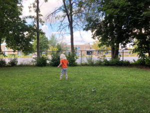 A toddler wearing an orange shirt walks away from the camera.