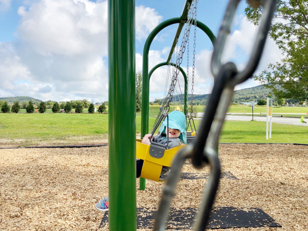 A toddler is wearing a blue hat, swinging on a yellow swing. The chains and poles of the swing set frame her.