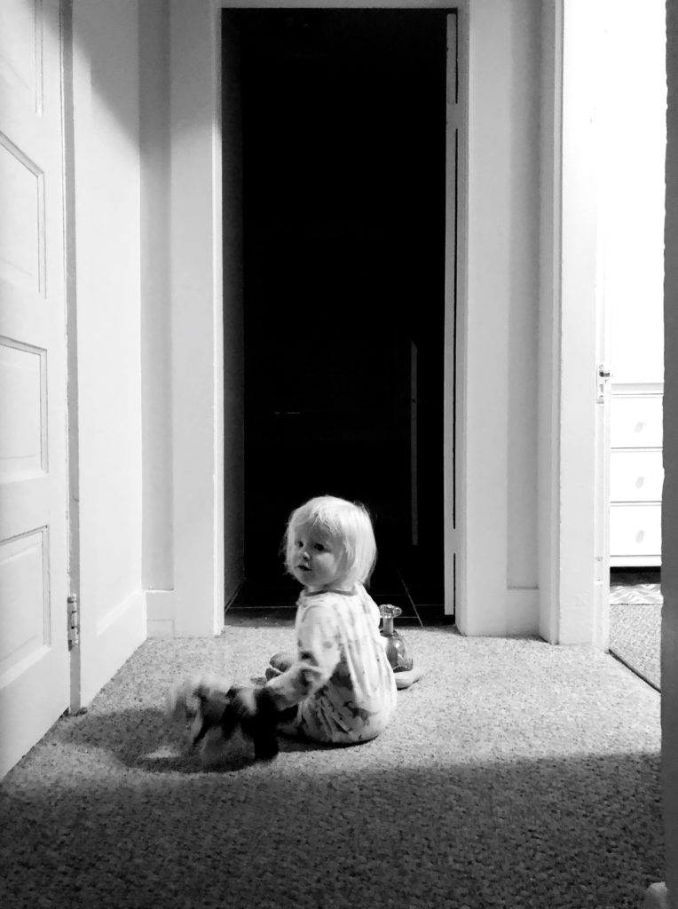 a toddler sits on the floor of a hallway in this black and white image. A stream of light comes from a door on the right.