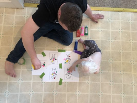 A man helps a toddler apply fingerpaint to paper taped to the floor.