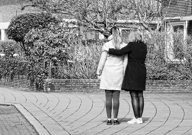 Two women stand facing away from the camera, arms around each other
