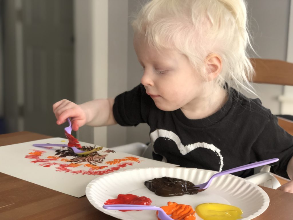 A toddler paints on a piece of paper using a fork.