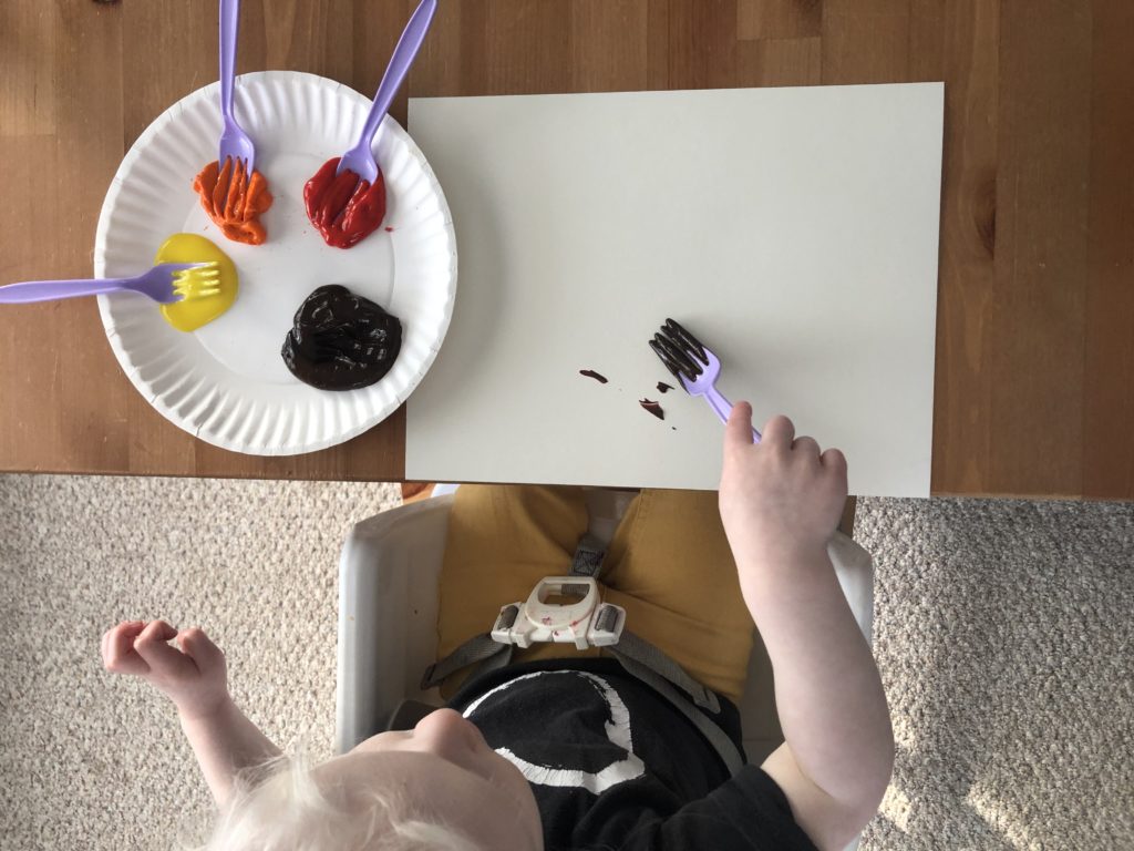 Viewed from above, a toddler uses a fork to paint on paper.