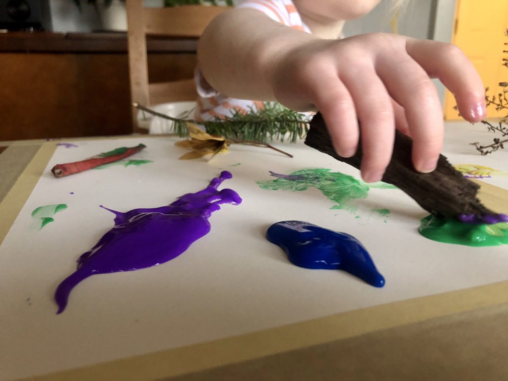 A toddler’s hand reaches towards the camera, dipping a piece of bark in green paint.