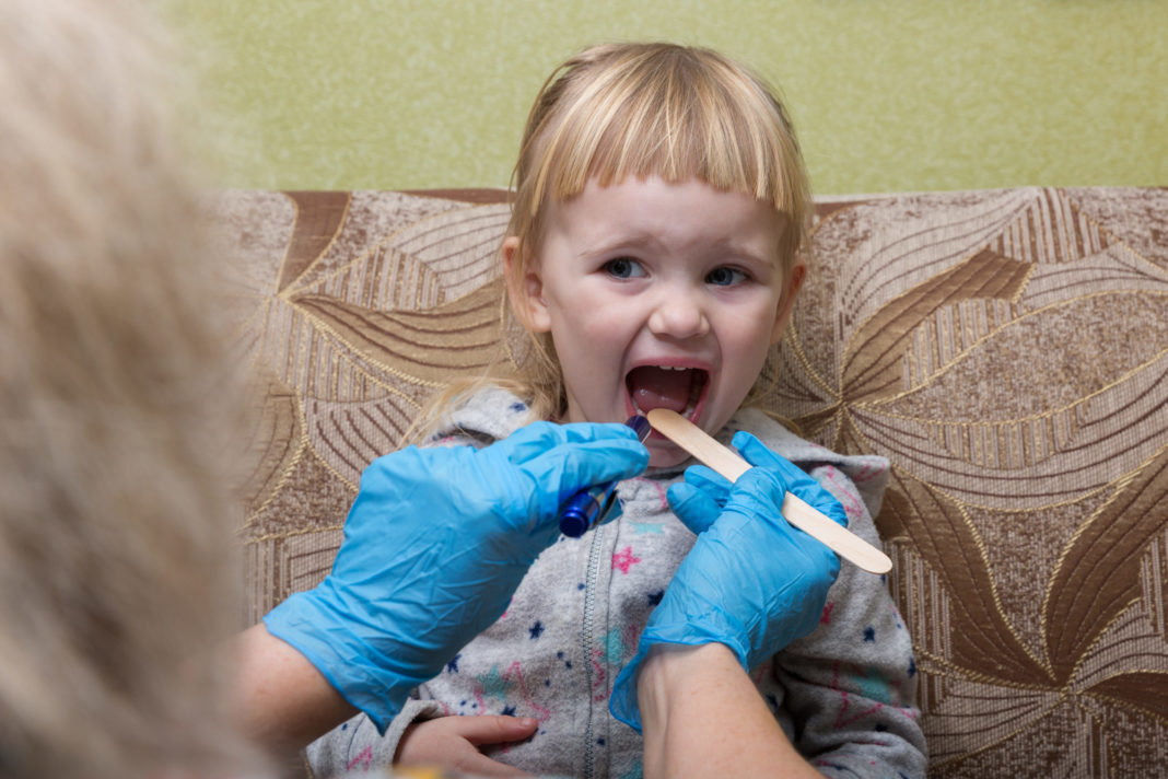 Tips for Managing Your Kid’s Sore Throat from a Pediatric Nurse Little girl with dirty blond hair and bangs sticks out her tongue for a tongue depressor to be used by a person wearing blue gloves.