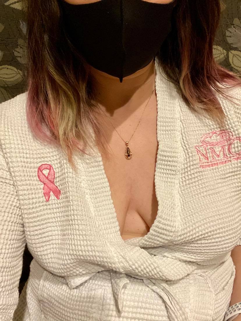 woman's torso, wearing a robe and covid mask. cleavage exposed. Breast cancer ribbon on robe.