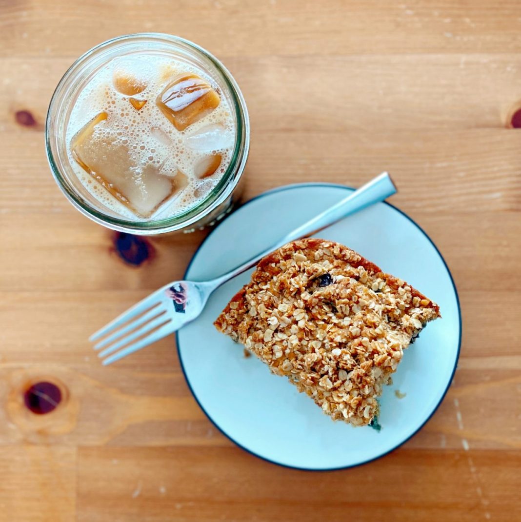 Viewed from above, an iced coffee next to a plate with oat milk blueberry muffin square