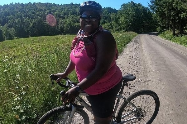A Black, femme, fat woman with a huge smile poses on a dirt road with a bicycle.