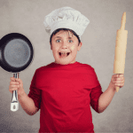 child in a red t shirt holding a rolling pin and a pan looking stressed