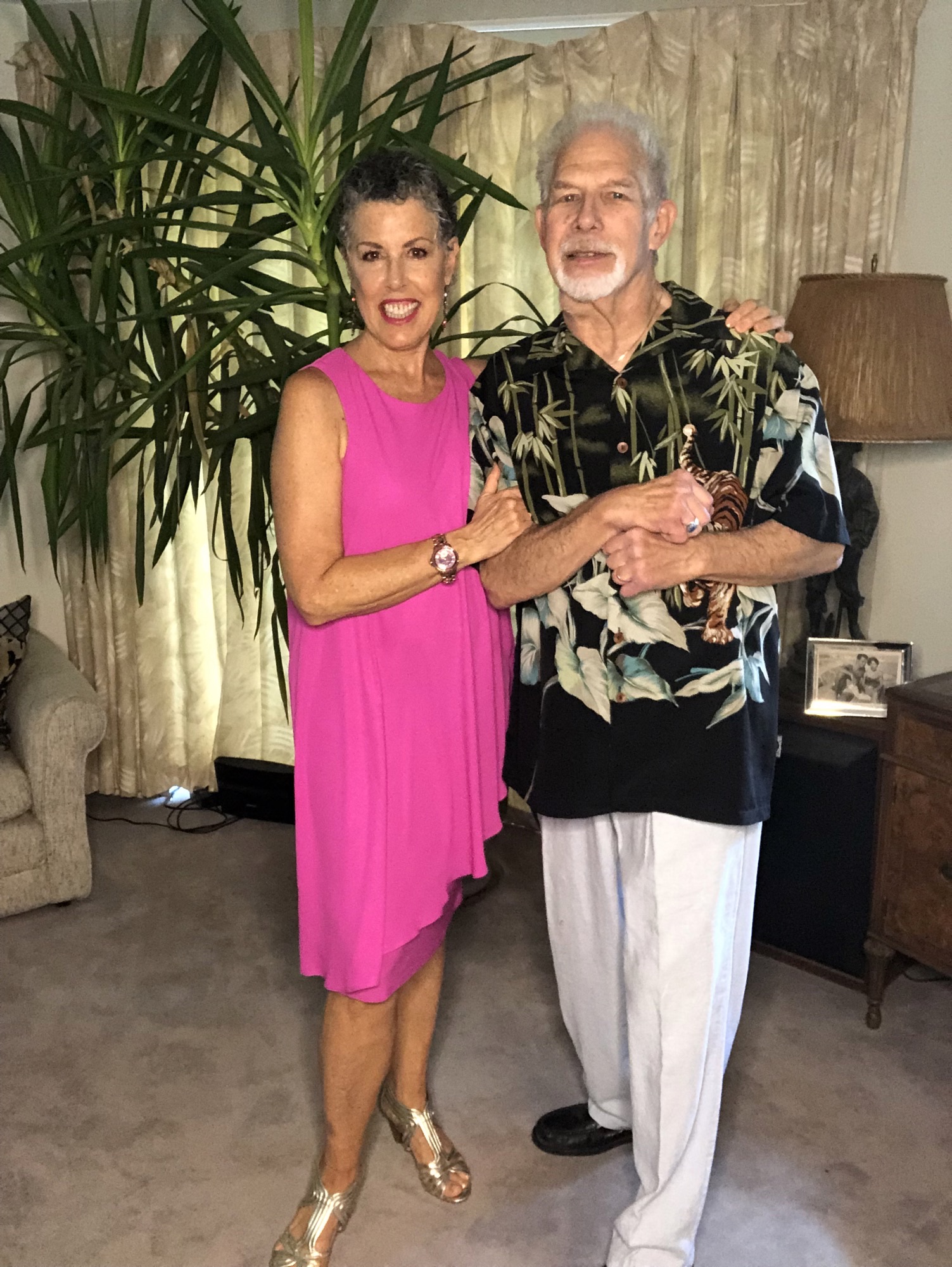 woman in a vibrant pink dress posing with her husband, who is wearing a shirt with a leaf print. 