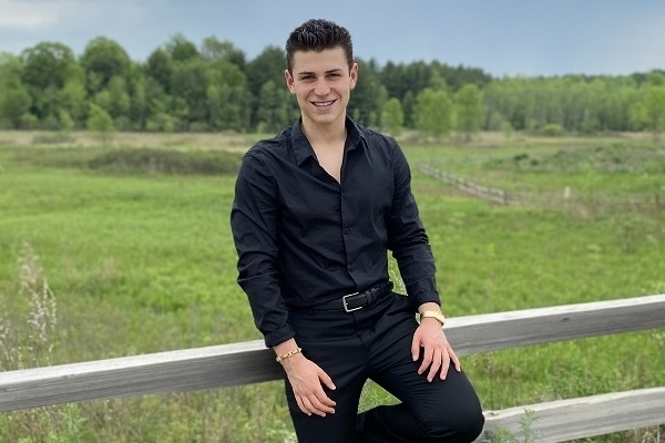 A young man dressed in a formal black suit leans casually against a wood rail fence. He is smiling.