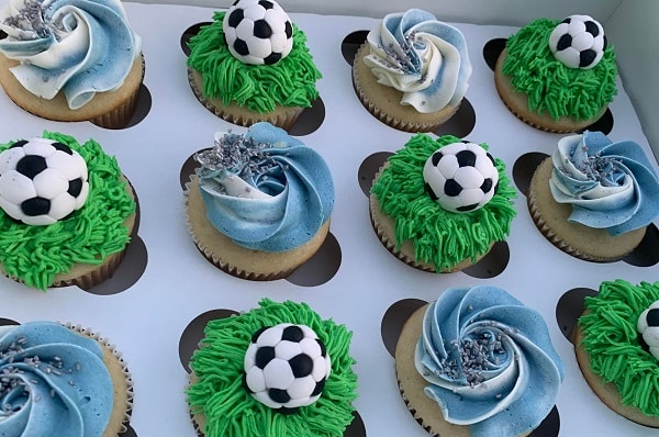A box of decorative blue and green cupcakes with soccer balls on them.