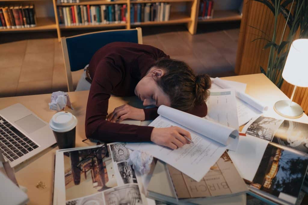 woman laying on desk full of papers, a to-go mug, crumbled paper, and her laptop.