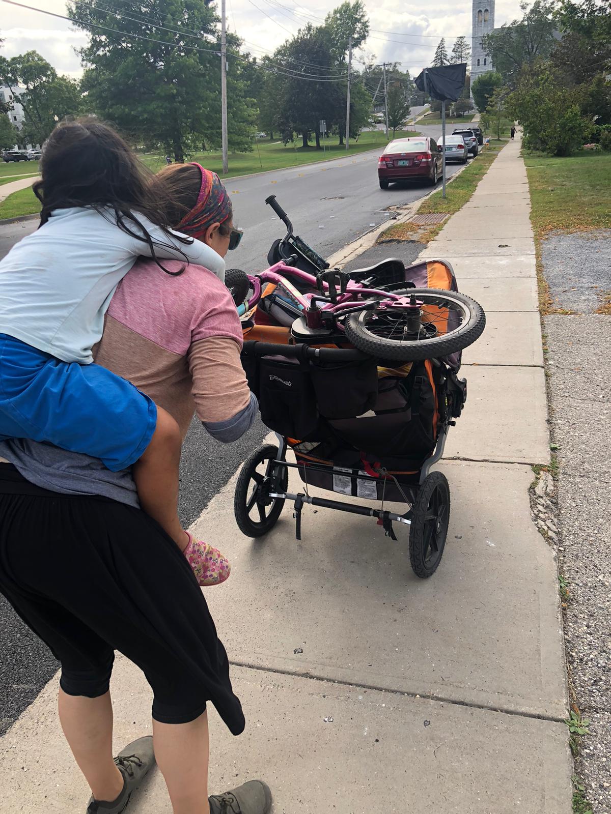 7ecf6478-852d-48ef-9dfe-50f583276892 Hasidic Jewish woman pushing a double stroller with a bike on top of the stroller while carrying a 4 year old on her back