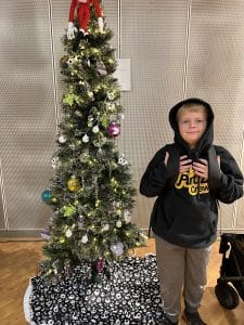 Boy in a hooded sweatshirt stands next to a Christmas tree