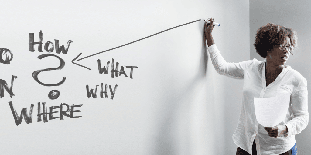 Woman standing in front of whiteboard with the words: How, What, Why, Where, and ?
