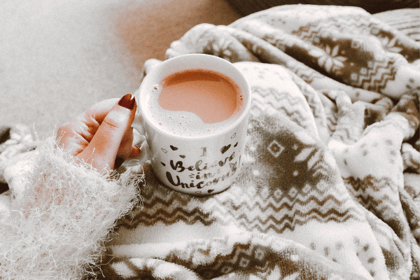 A woman's hand holds a mug of hot coffee on top of a snowflake-patterned throw.