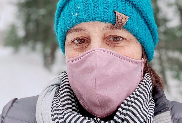 A woman dressed in a turquoise hat, pink mask, and striped neck wrap outside in winter snow.