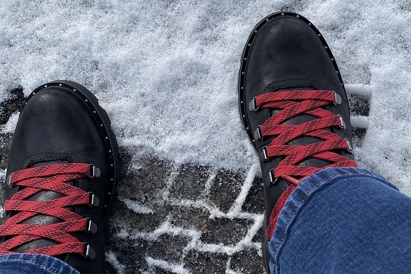 Black leather boots with red laces on snowy pavement over a tire track.