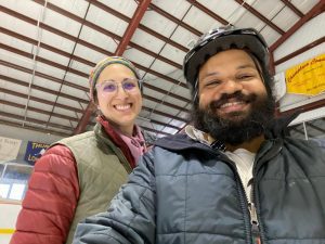 man with beard in foreground and woman slightly behind him. Both are smiling