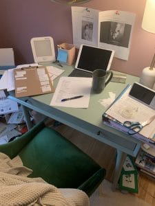 A green desk in a pink room. On the desktop are cut out pieces of paper, a print out, an iPad, a coffee mug, folders, and a baby monitor.