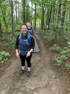 A white woman stands on a trail path with a baby in a carrier on her back. She wears black leggings and a black t-shirt, a blue fleece vest, and hiking shoes. She smiles.