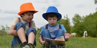 The One Simple Thing I Do to Get My Kids to Read Two little boys wearing hats are sitting in a field of grass. One is reading a book to the other one.