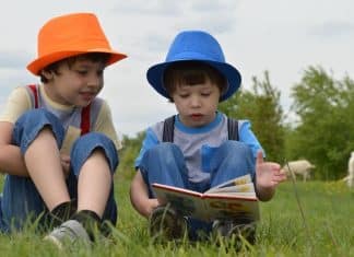 Two little boys wearing hats are sitting in a field of grass. One is reading a book to the other one.