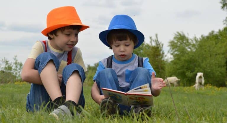 Two little boys wearing hats are sitting in a field of grass. One is reading a book to the other one.
