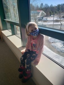 A young girl with a mask sitting in front of a big window overlooking a snowy walkway