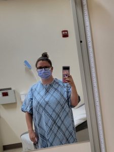Woman in hospital gown and mask takes selfie in mirror inside doctors office room