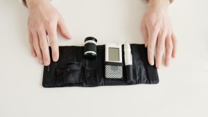 Two white hands hold open a diabetes testing kit. It includes a capsule of blood testers, the reader, and finger stick. It lays on a white surface.