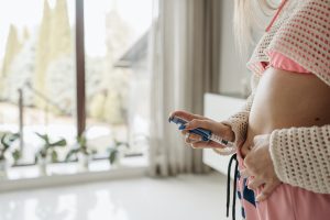 A white woman with long purple fingernails pinches a bit of her belly with one hand and holds a syringe against it with the other. She’s wearing pink bottoms and a pink bra. A bathtub is behind her and open windows look out.
