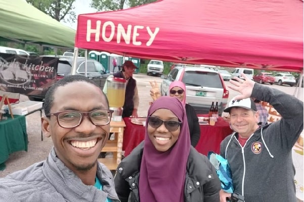 A Black man and woman, and a white man, wave by a tent labeled "honey" at the farmers market.