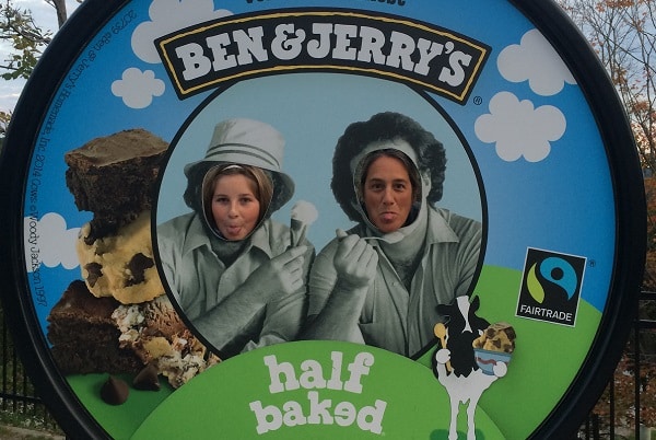 A woman and a girl stick out their tongues as they look thruogh a giant Ben & Jerry's ice cream lid.