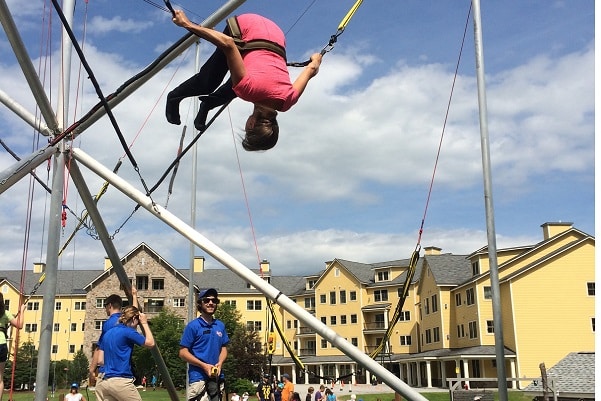 A woman does a flip assisted by rubber bands and a trampoline, against a mountain backdrop.