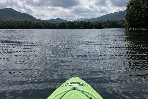 The stern of a green kayak is in the foreground, leading to calm water and a mountain view.