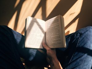 A person reads a book cross legged on a wood floor. Sunlight casts shadows on the book through the window.