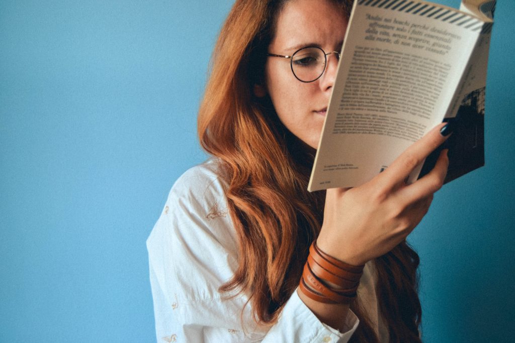 A white woman with red hair and glasses read a book in front of a blue wall.