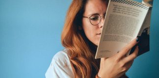 Jessi Klein’s Book, I’ll Show Myself Out, Is Everything I Needed A white woman with red hair and glasses read a book in front of a blue wall.