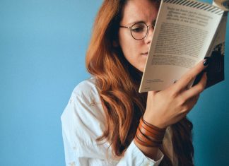 Jessi Klein’s Book, I’ll Show Myself Out, Is Everything I Needed A white woman with red hair and glasses read a book in front of a blue wall.