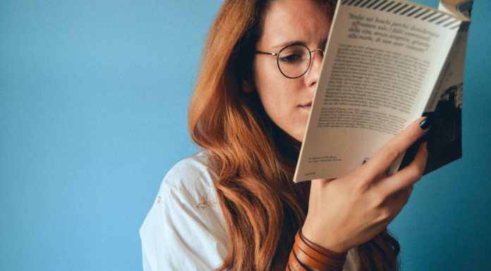 Jessi Klein’s Book, I’ll Show Myself Out, Is Everything I Needed A white woman with red hair and glasses read a book in front of a blue wall.