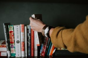 The hand of a white woman reaches for a book from a full shelf. Her sleeve is cuffed and she wears a watch and many bracelets on her wrist.