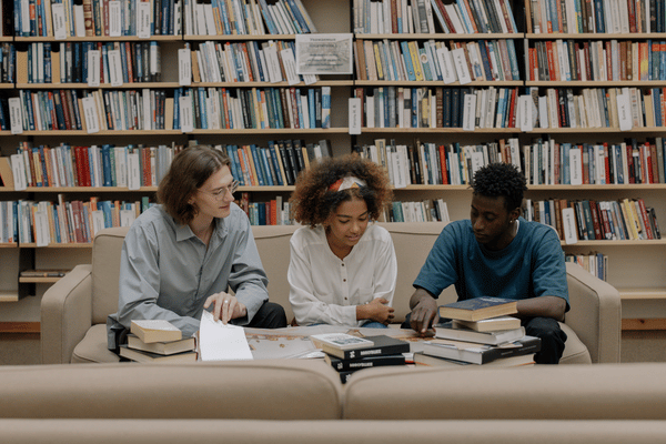 Students sit at a table in front of stacks of books.
