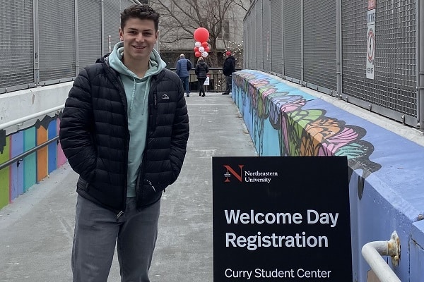 A boy stands in front of a university welcome sign.